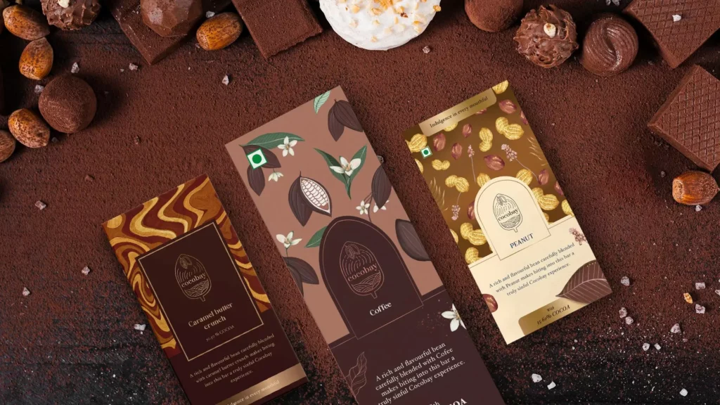Flat lay of Caramel Whirl, Cocoa, and Peanut flavored chocolate bars surrounded by assorted chocolates and cocoa powder on a dark surface.